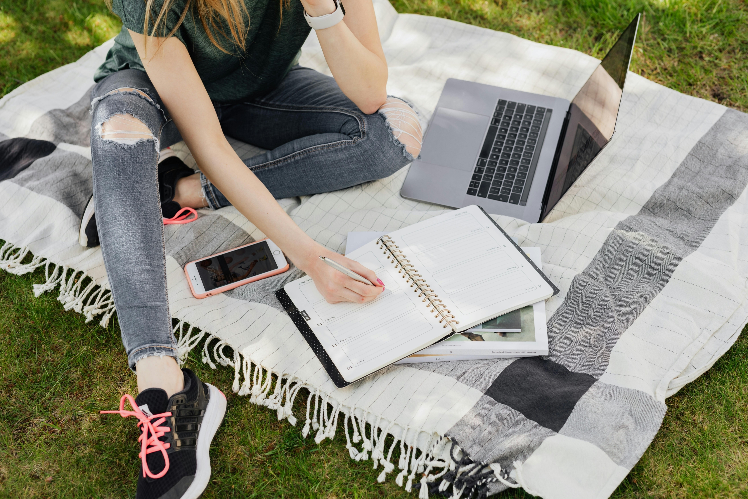 A student working on a summer course on a picnic blanket, with her phone, computer and a notebook.